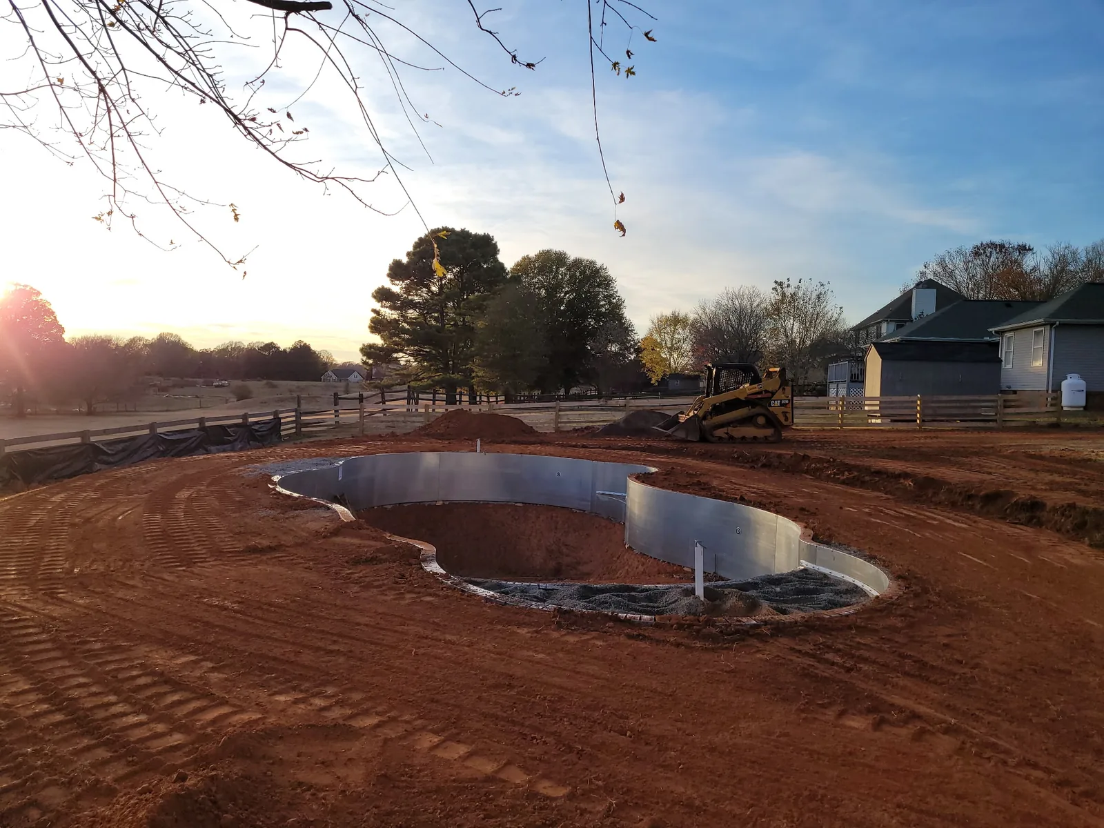 A freeform pool excavation with a skid steer working the backfill at sunset — Little Acres Grading